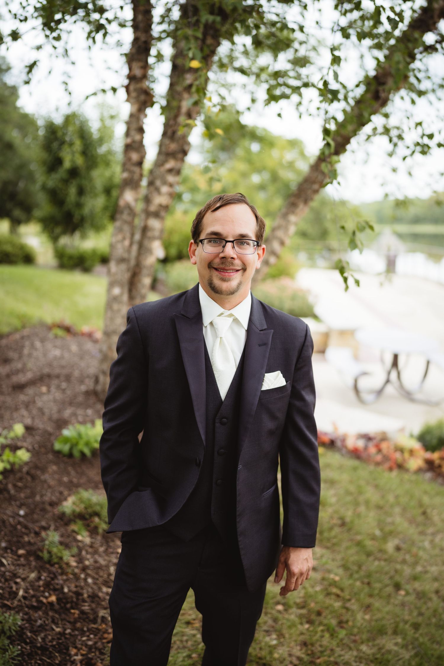 A person in a formal black suit and white shirt stands outdoors near trees in a natural setting.