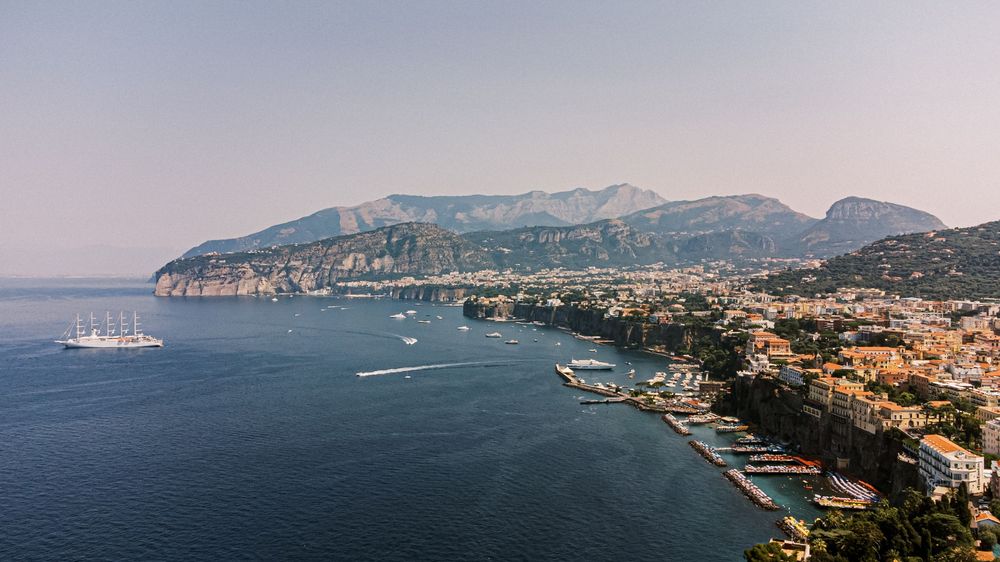 A panoramic view of the Mediterranean coast shows boats in a harbor with mountains and buildings along the shoreline.