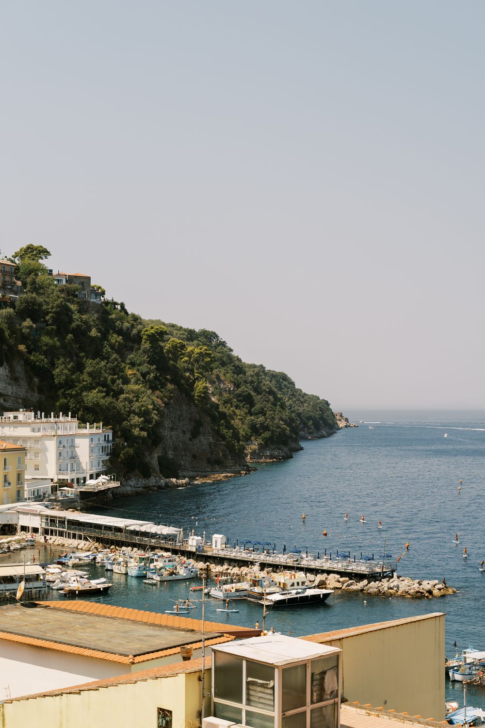 Coastal marina with boats docked along pier next to tree-covered cliffs and buildings overlooking blue Mediterranean waters.