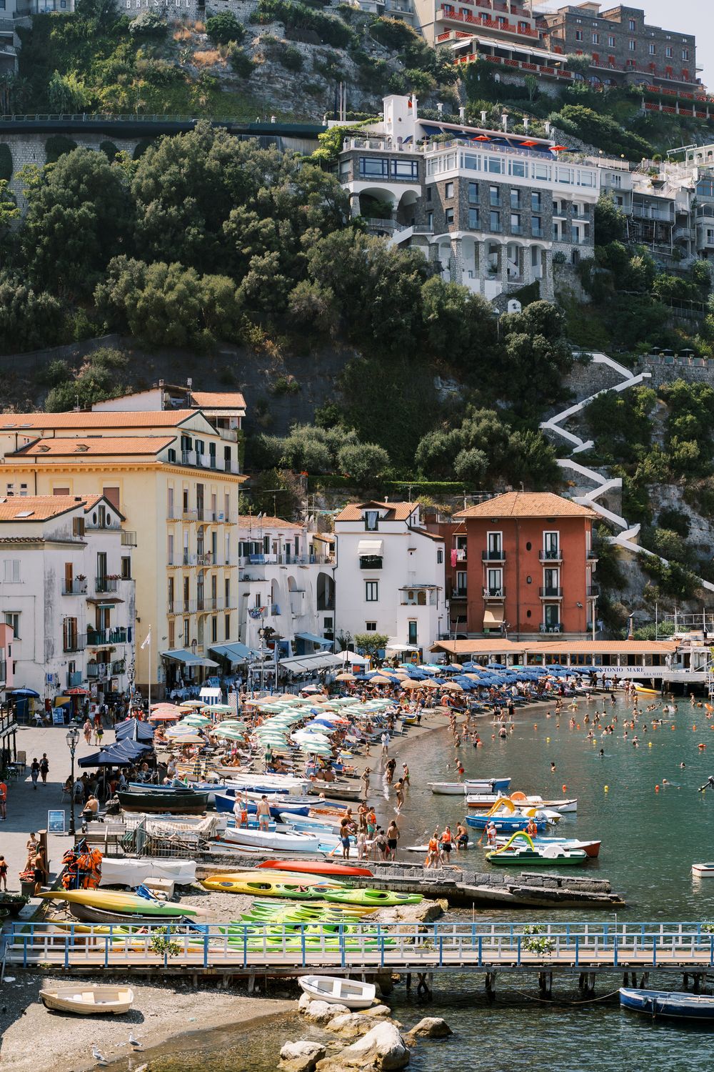 Colorful boats line the beach and pier at a scenic Mediterranean coastal village with hillside buildings.