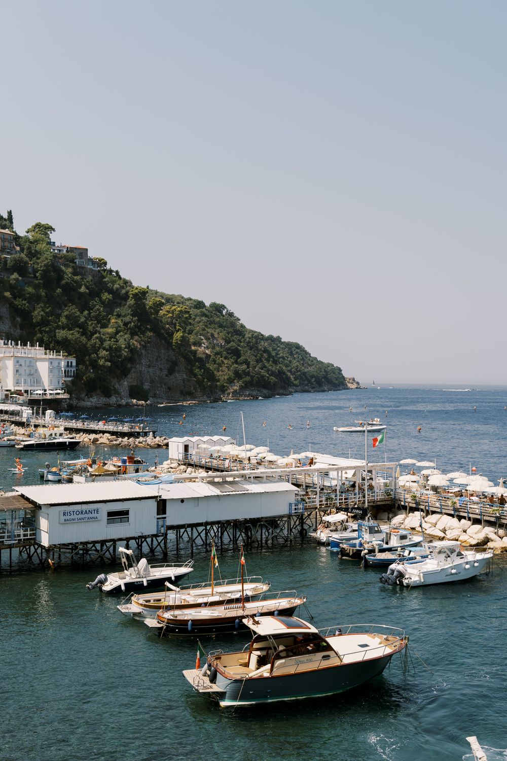 Boats docked at a scenic Mediterranean pier with white umbrellas and coastline visible in blue waters.