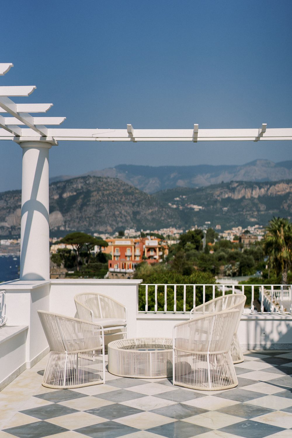 White rattan chairs arranged on a tiled terrace overlooking Mediterranean mountains and coastal buildings.