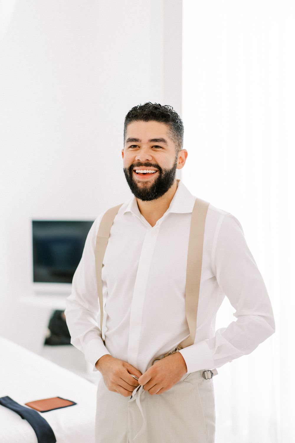 Bearded man in white shirt and suspenders smiling in a bright room.