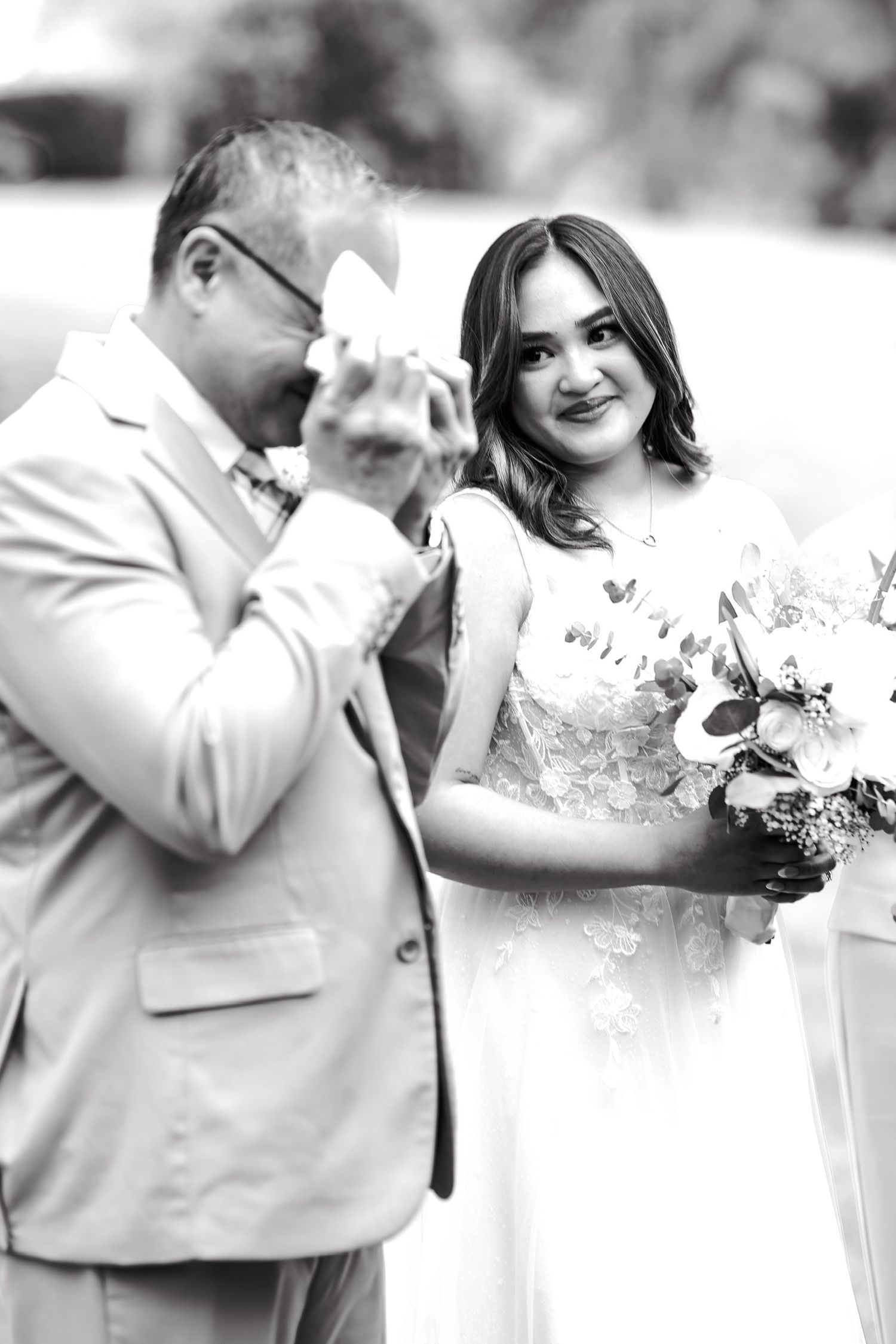 Black and white wedding photo showing an emotional moment between a suited man wiping tears and smiling bride holding bouquet.