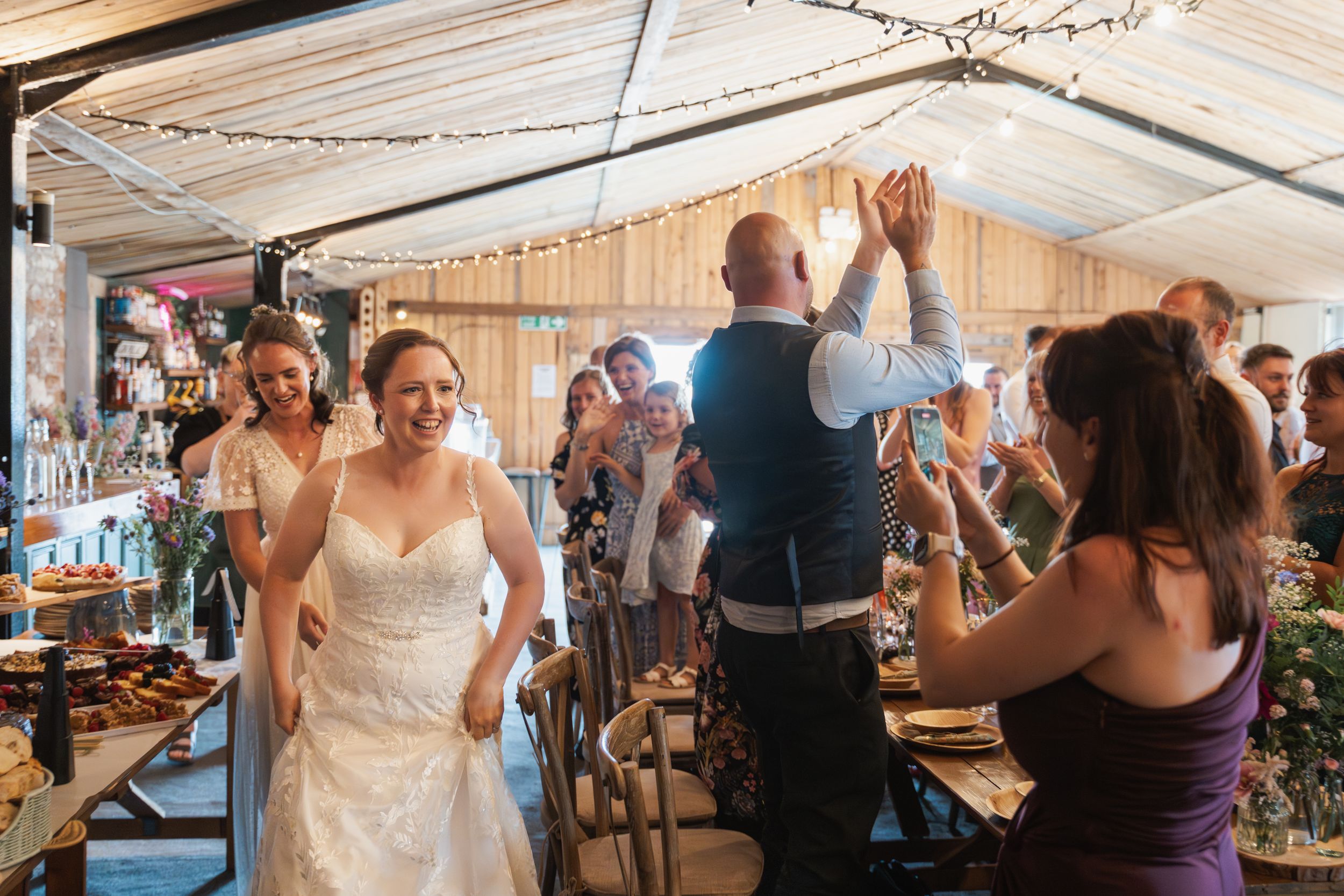 Wedding guests celebrate with dancing and cheering inside a rustic barn venue decorated with string lights.