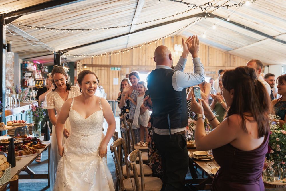 Wedding guests celebrate with dancing and cheering inside a rustic barn venue decorated with string lights.