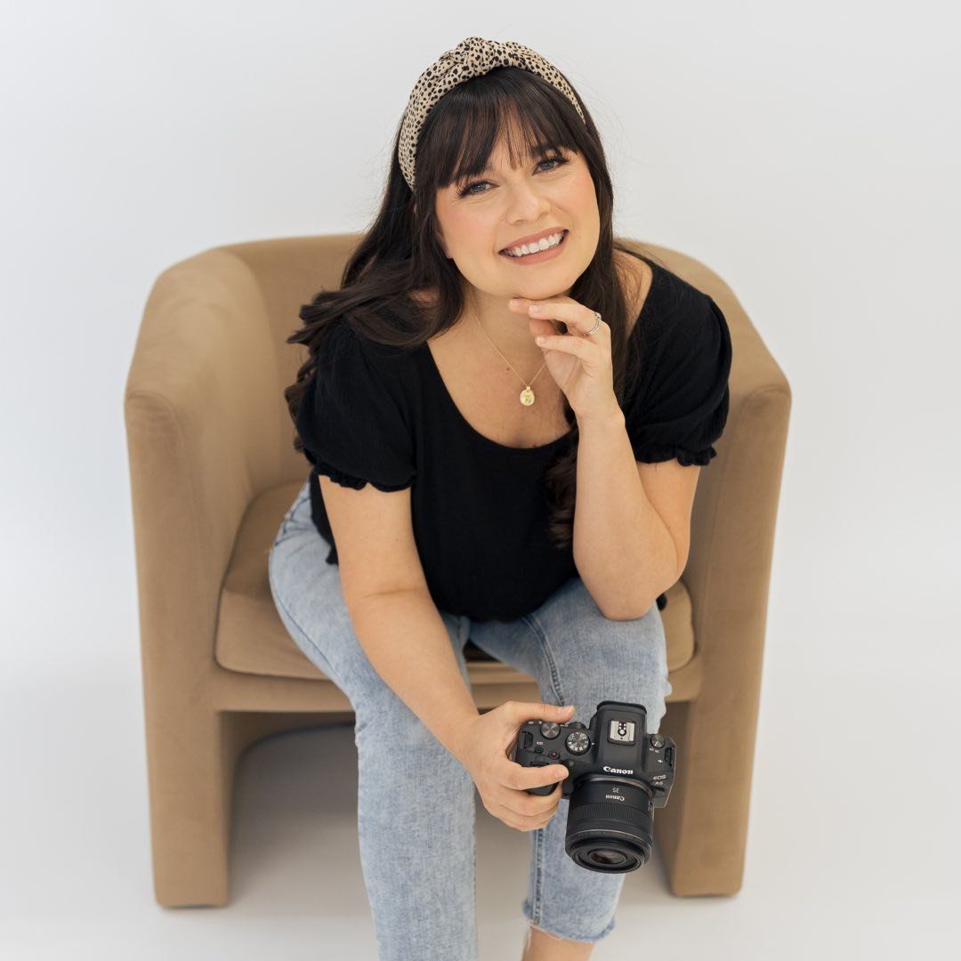 Smiling female photographer sits in a chair holding a camera with a leopard print headband.