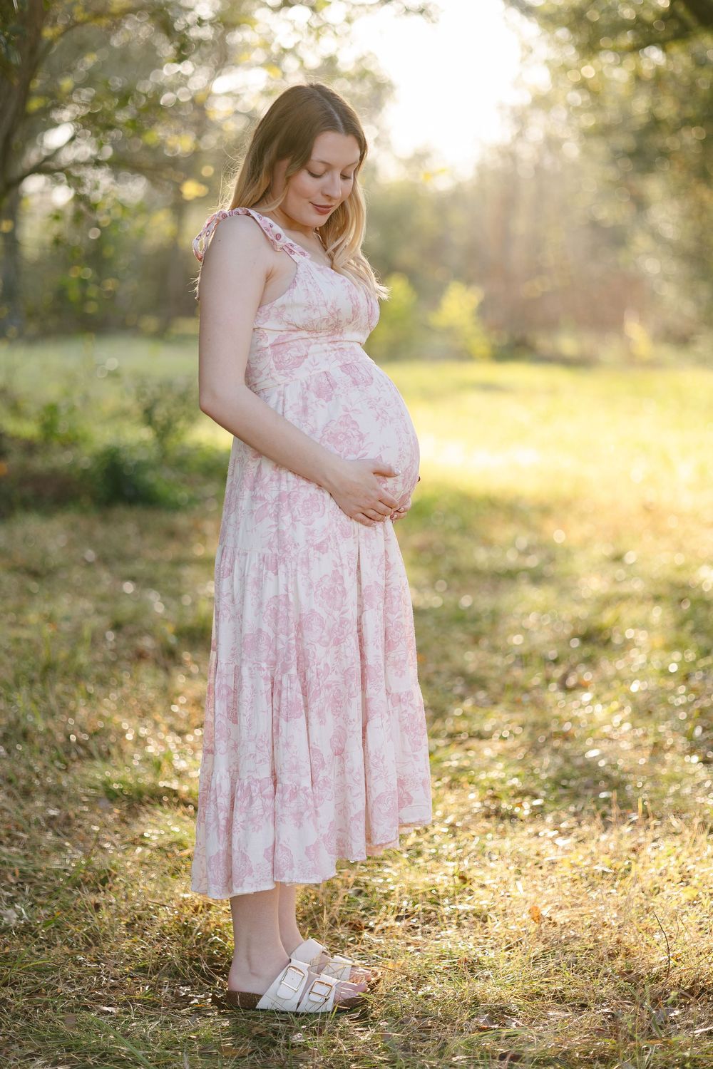 A pregnant woman in a pink floral dress stands in a sunlit outdoor setting during golden hour.