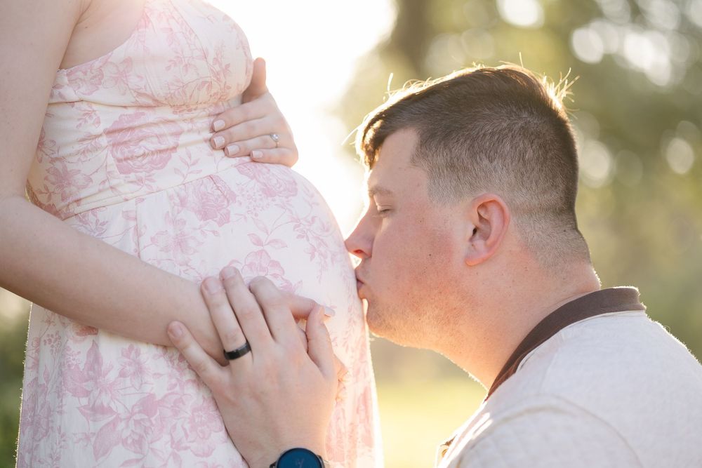 A tender maternity photo showing a gentle kiss on a pregnant belly during golden hour lighting outdoors.