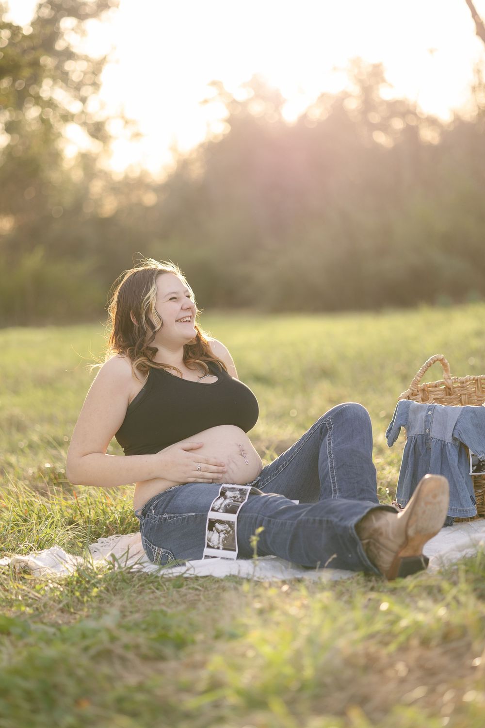 Pregnant woman sitting on blanket in golden sunlight, smiling while holding ultrasound photos on her baby bump outdoors.