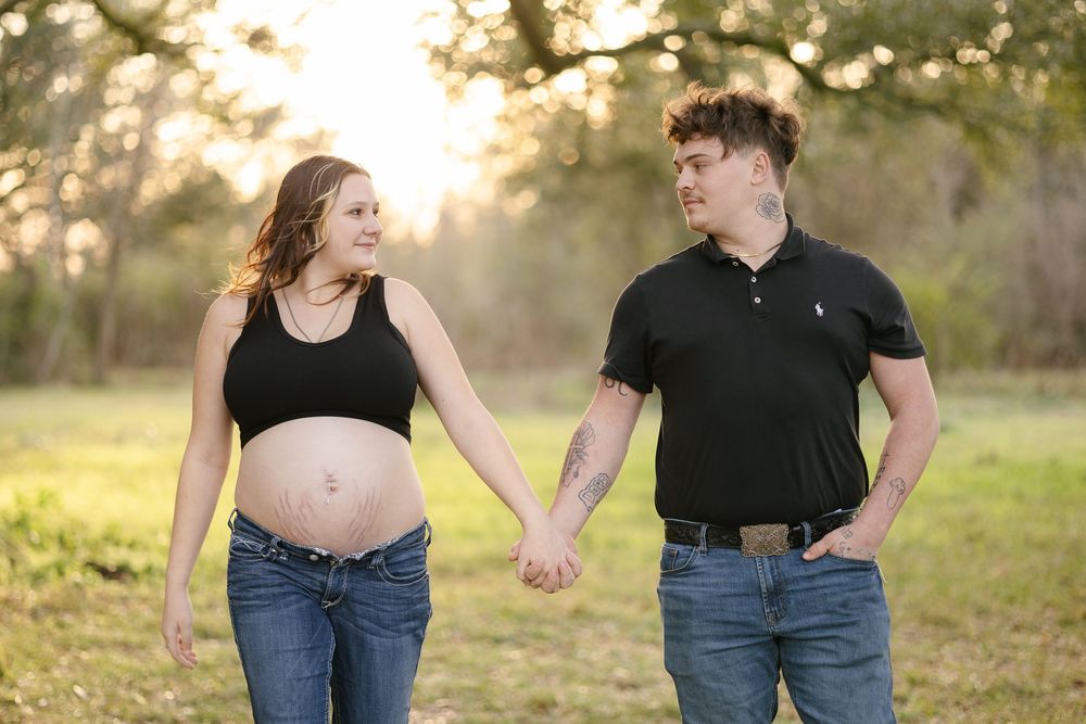 Pregnant couple holding hands in a sunlit park, woman showing baby bump in black crop top and jeans.