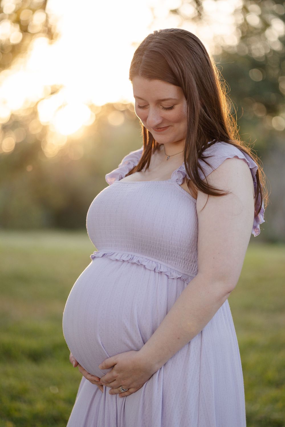 Expectant mother in flowing white dress cradles her baby bump during golden hour maternity photoshoot in a grassy field.