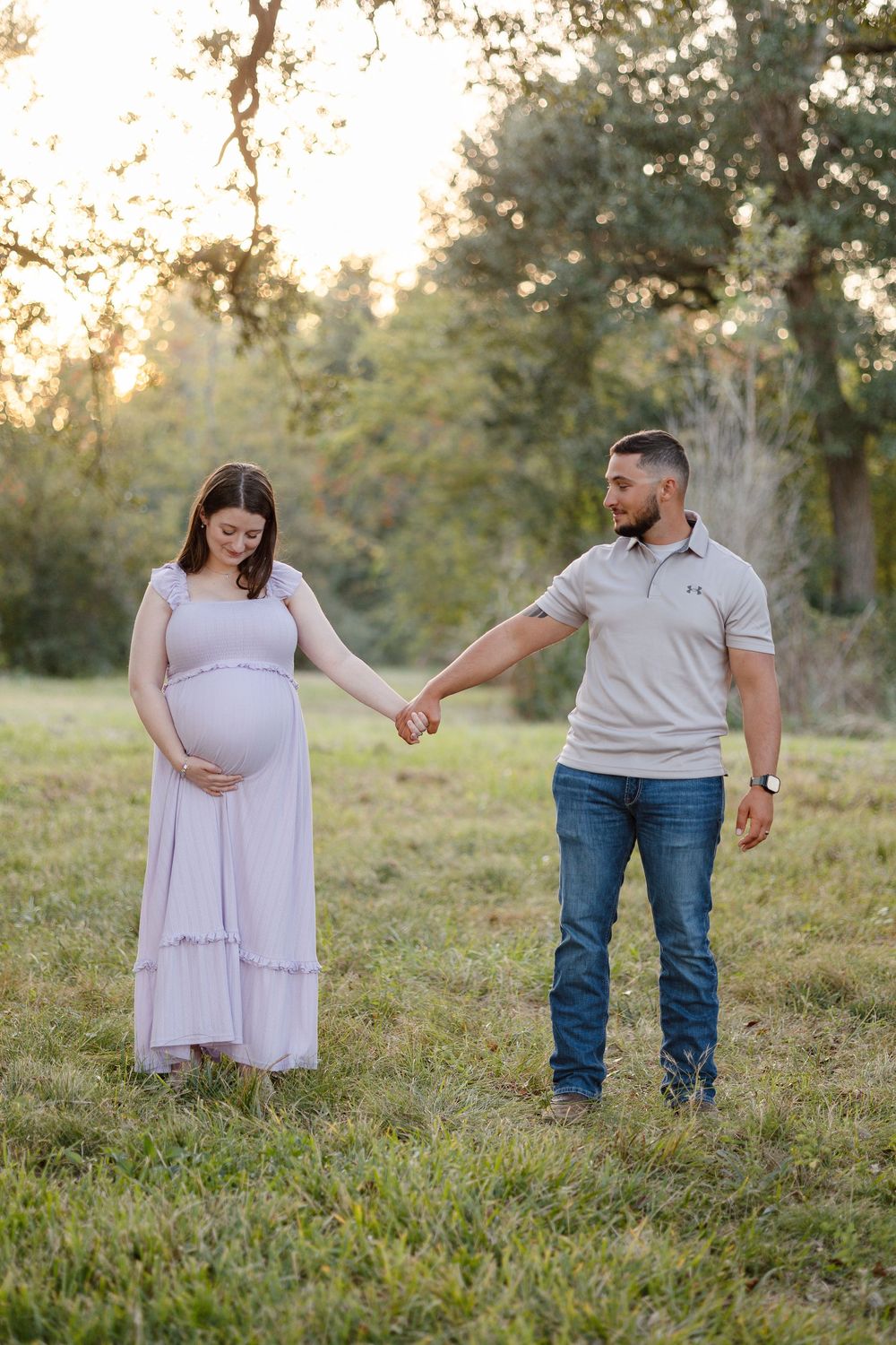A couple holds hands in a grassy field at sunset while expecting their first child.