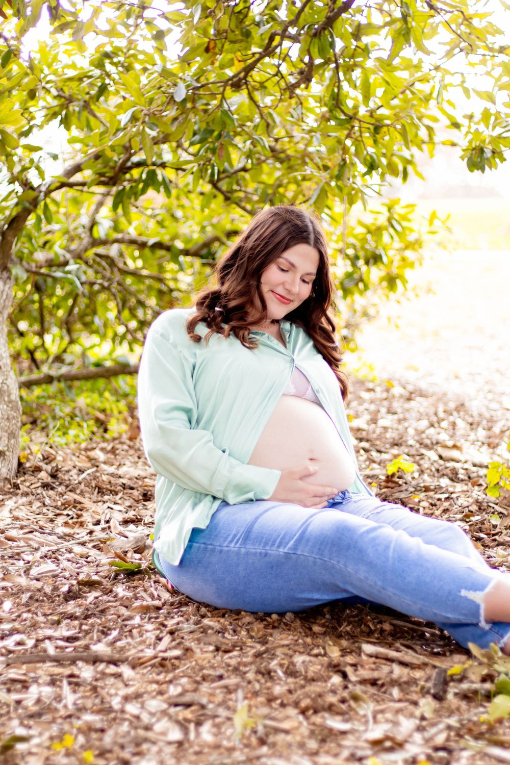 Pregnant woman sitting under a tree, cradling her baby bump, wearing a mint cardigan and jeans outdoors.