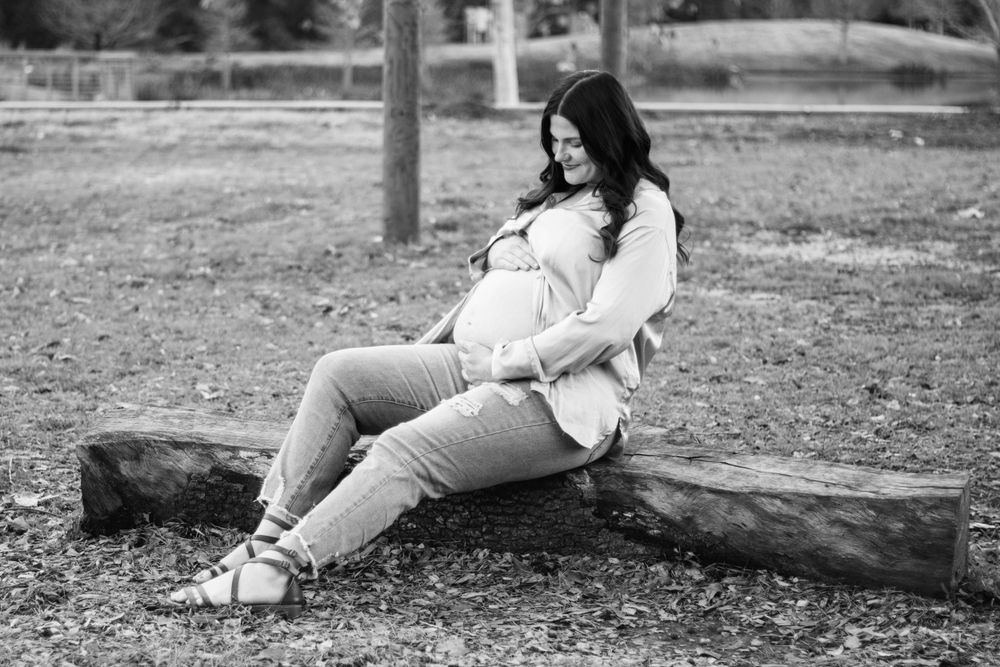Black and white maternity photo of a pregnant woman sitting on a log in a park, cradling her baby bump.