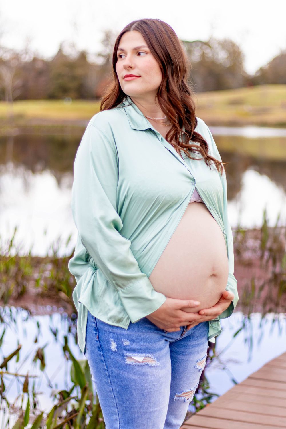 Pregnant woman in mint shirt and jeans cradling baby bump outdoors near a serene pond.