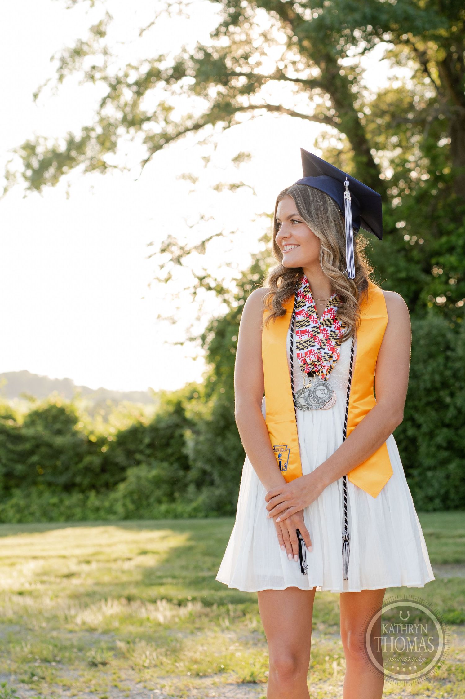 Graduate in white dress and academic regalia poses thoughtfully in golden sunset light.