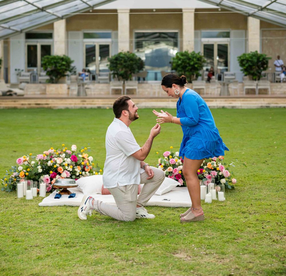 A proposal scene on a lawn with floral arrangements and a columned building in the background.