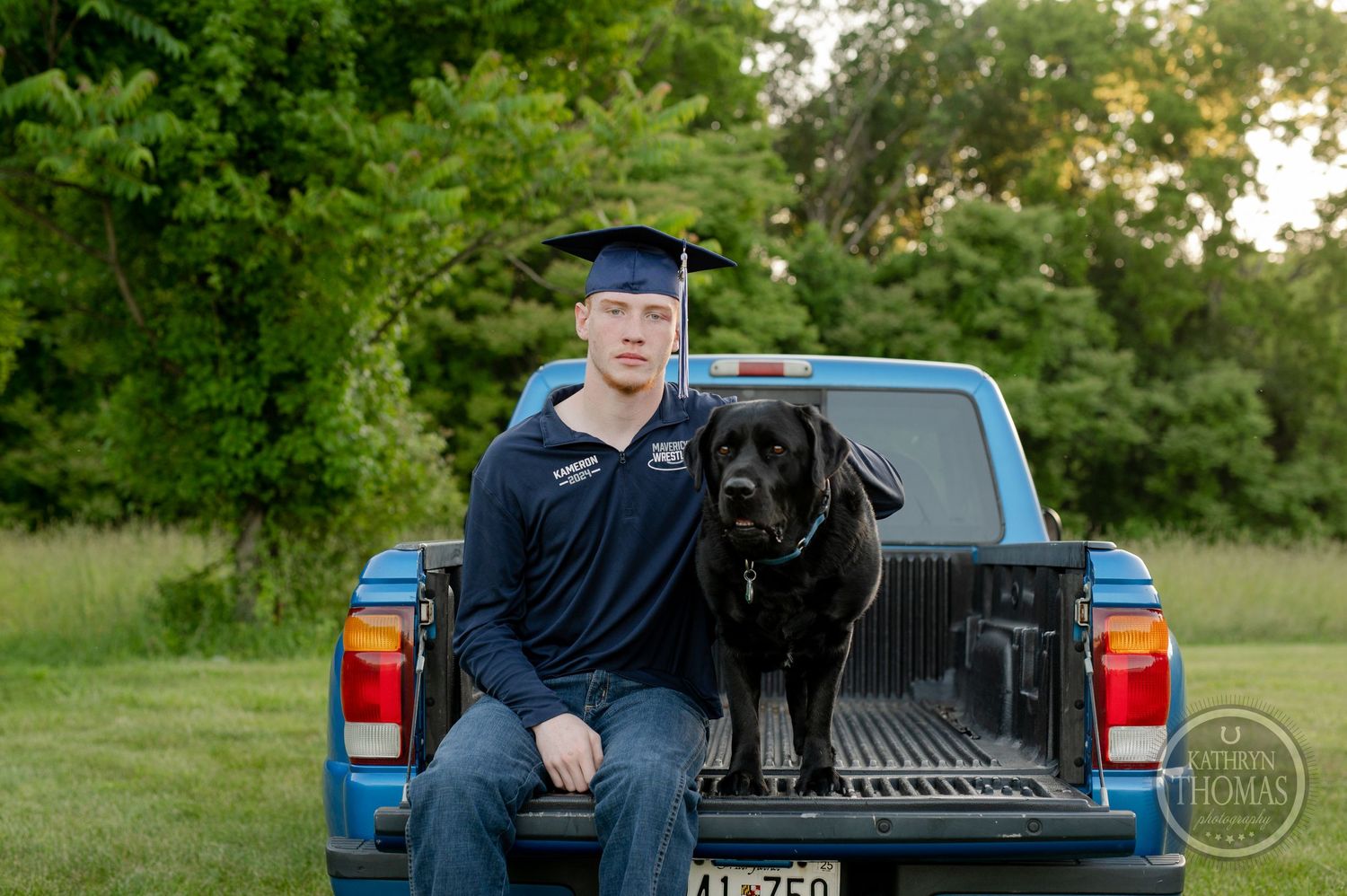 Graduate in navy attire sits with black dog in blue pickup truck bed.