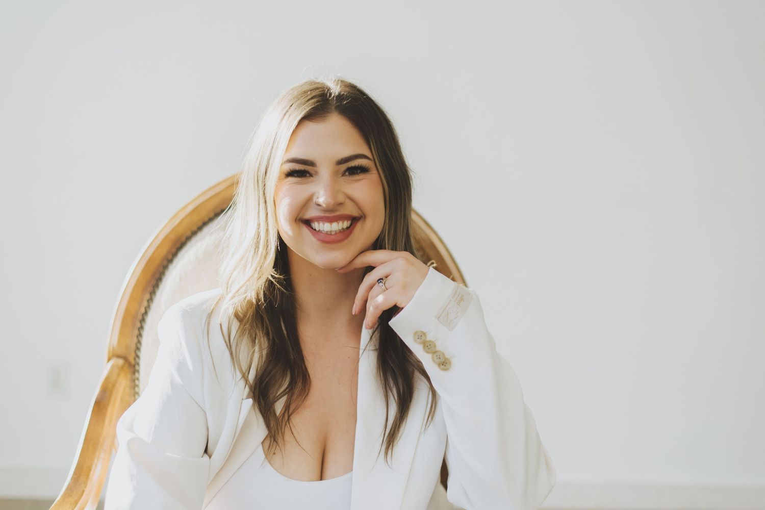 A woman with long brown hair wearing a white blazer smiles warmly while sitting in a wooden chair.