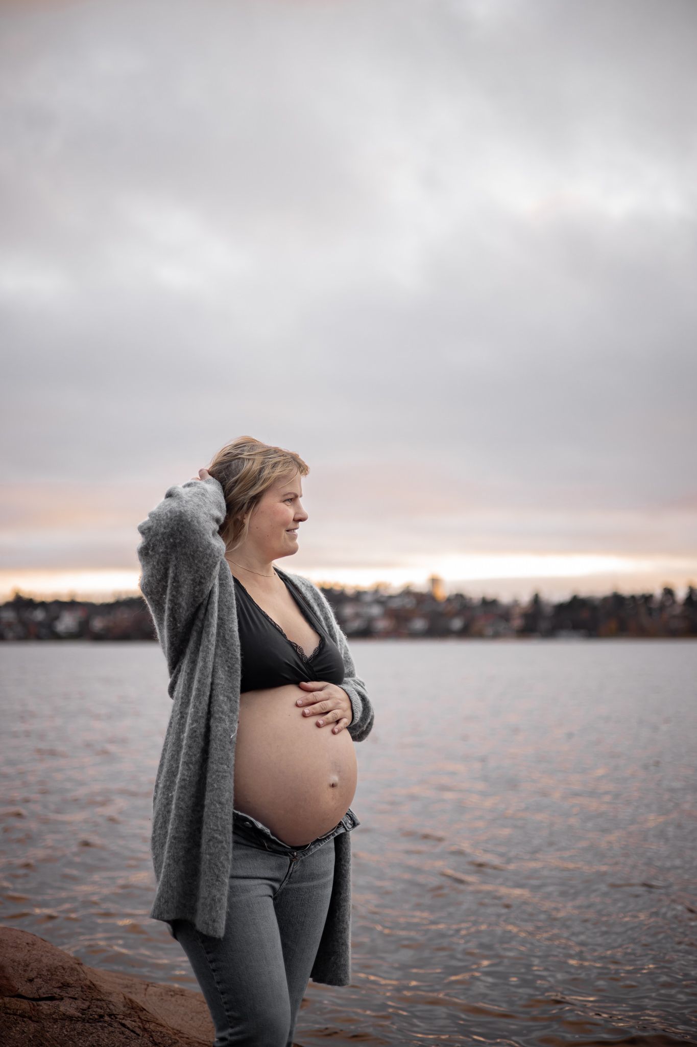 An expectant mother in a gray cardigan stands by a calm lake at sunset with cloudy skies overhead.