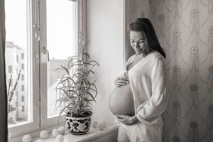 A black and white photograph of an expectant mother in a white sweater standing by a window with a potted plant.