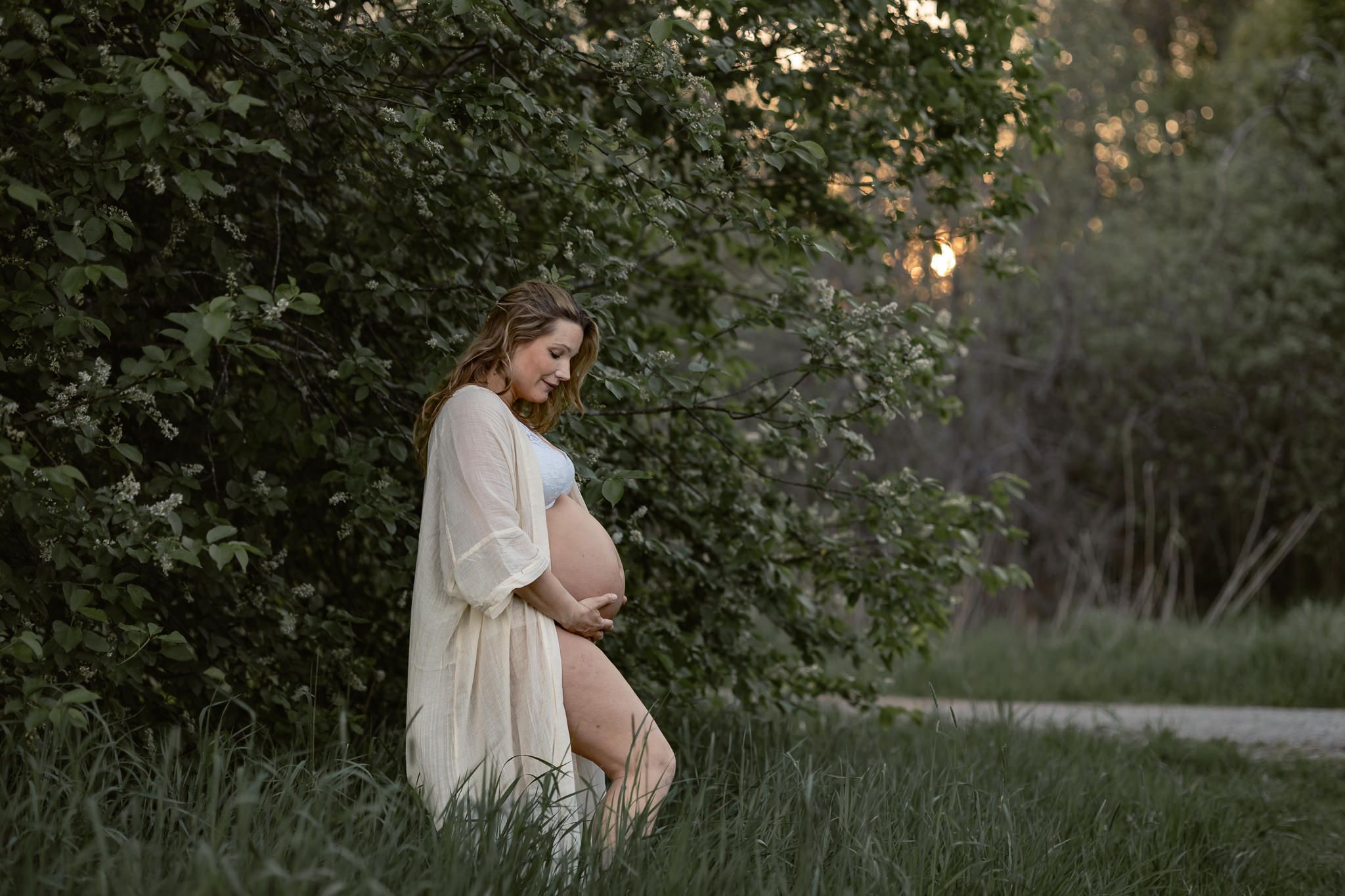 An expectant mother in a flowing white dress stands peacefully among lush greenery during golden hour.