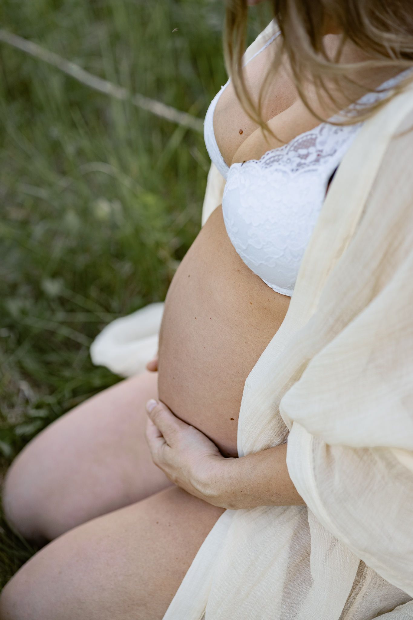 A pregnant woman in white lace bralette and flowing robe cradles her belly while sitting on grass.