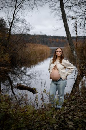 A pregnant figure stands by a serene river on an autumn day wearing a cream sweater and blue jeans.