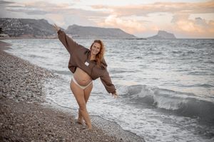 A joyful person dances on a rocky beach at sunset with mountains and ocean waves in the background.