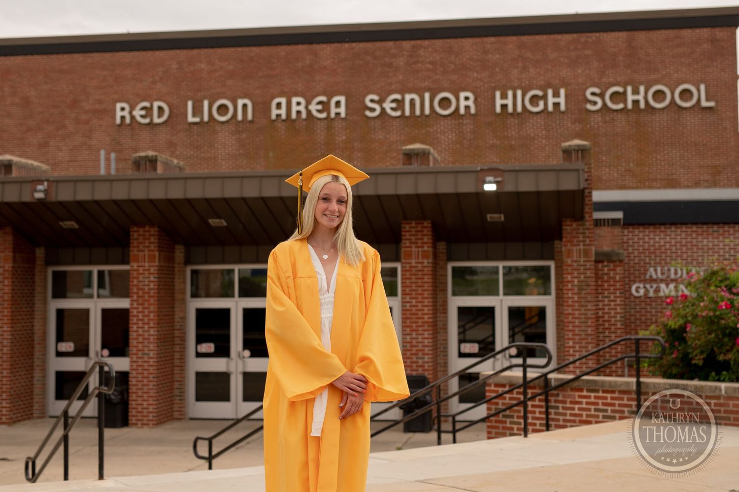 Graduate in bright yellow cap and gown poses in front of Red Lion Area Senior High School.