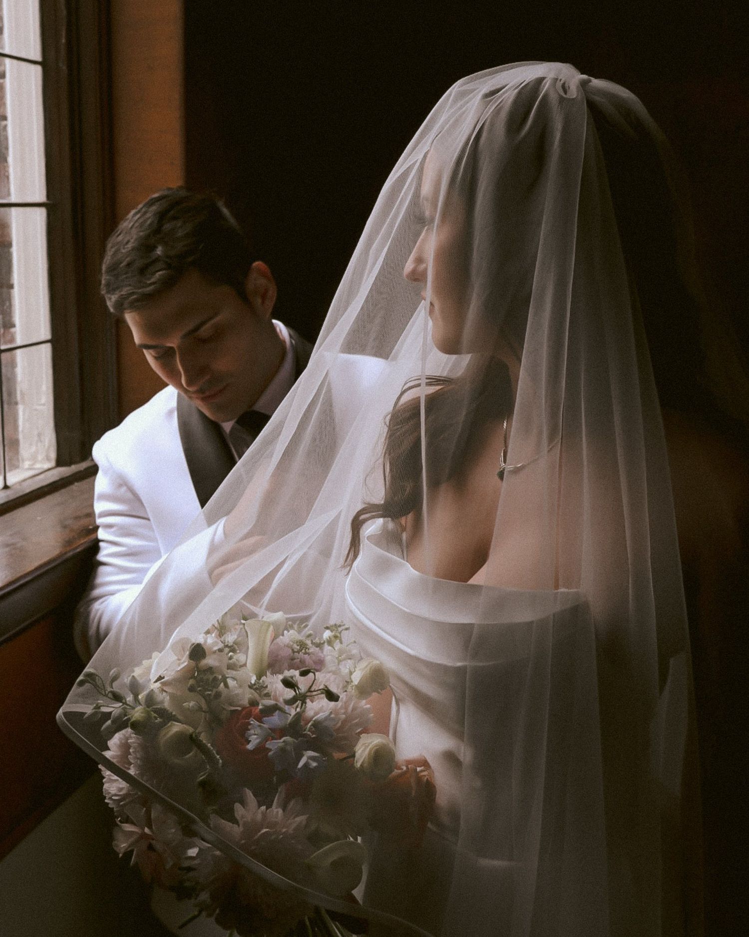 Moody portrait of couple by window with bride in veil holding bouquet and groom in white jacket.