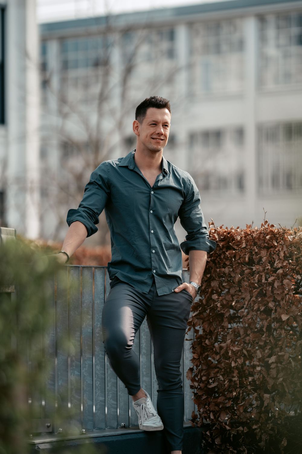 Person in casual attire poses casually on outdoor railing near hedges.