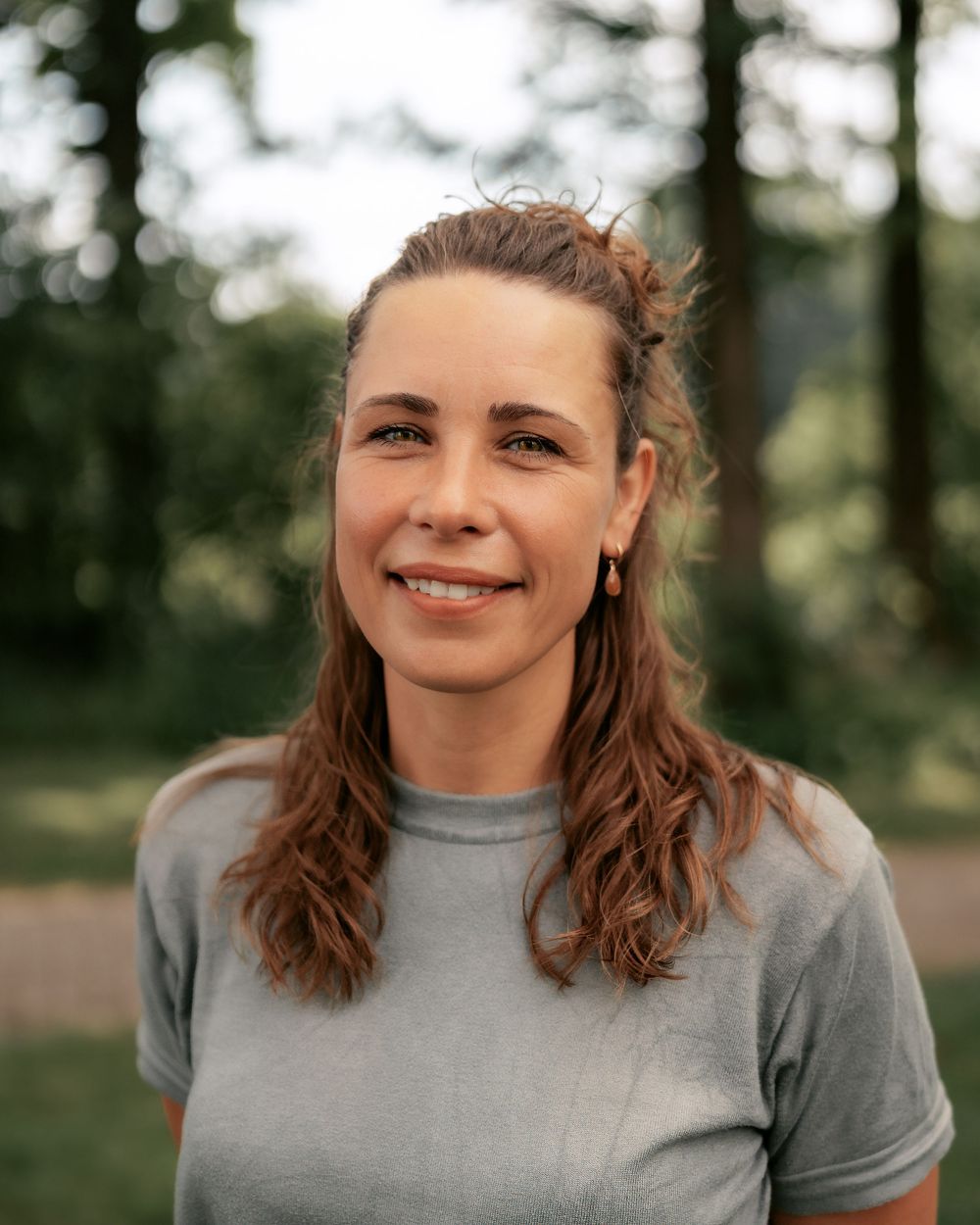 A person with wavy brown hair and half-up hairstyle smiles warmly while wearing a gray t-shirt outdoors.