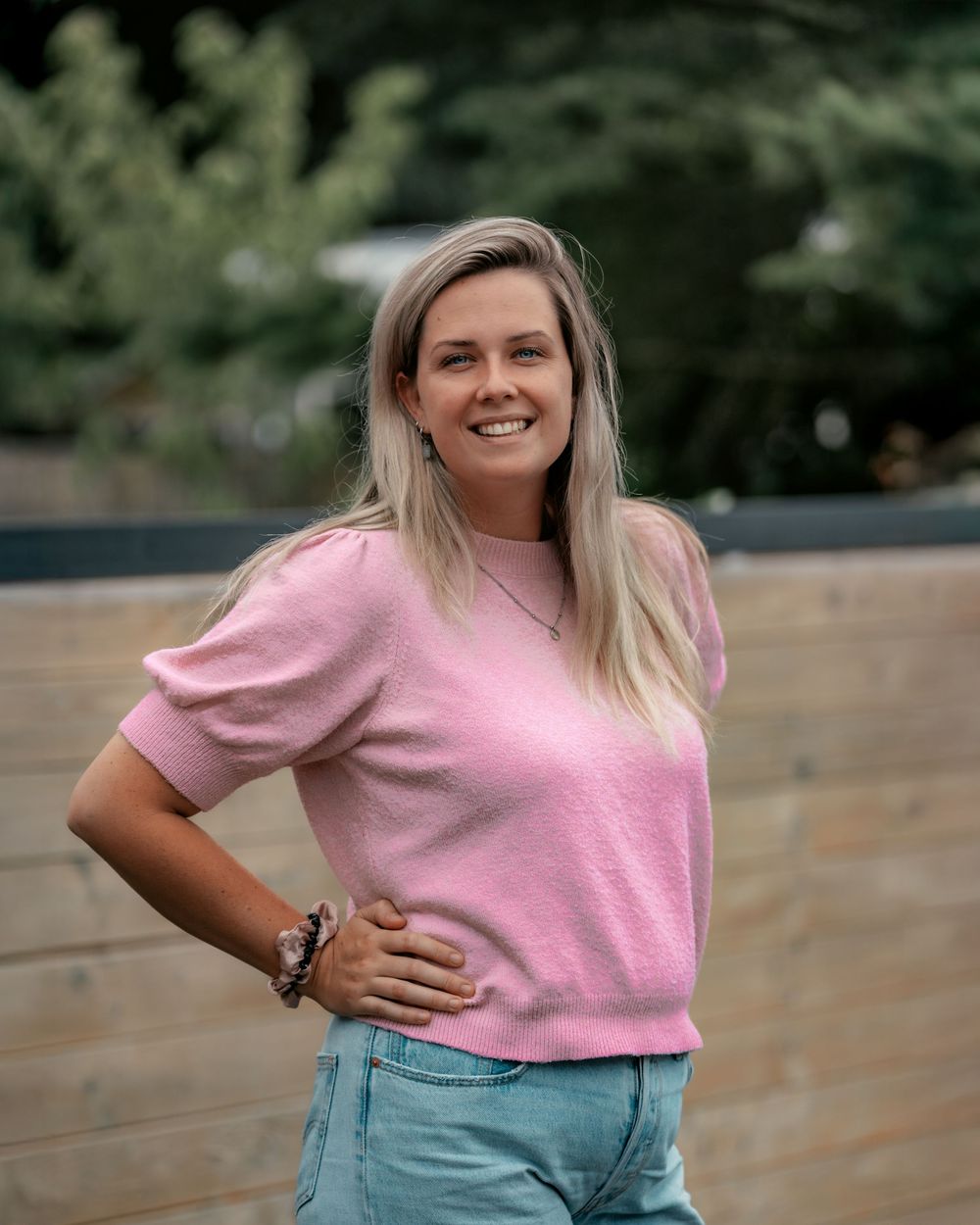 Person in pink sweater and light jeans leaning casually against outdoor wall with natural backdrop.