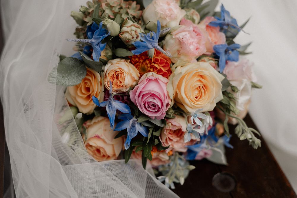 Wedding bouquet featuring peach roses, pink blooms and blue accents with delicate greenery.