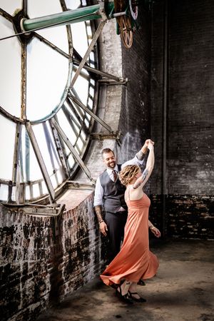 A dancing couple in formal attire perform in front of a large vintage clock face inside an industrial brick space.