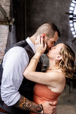 A romantic couple shares a passionate kiss in an industrial setting with brick walls and a circular window.