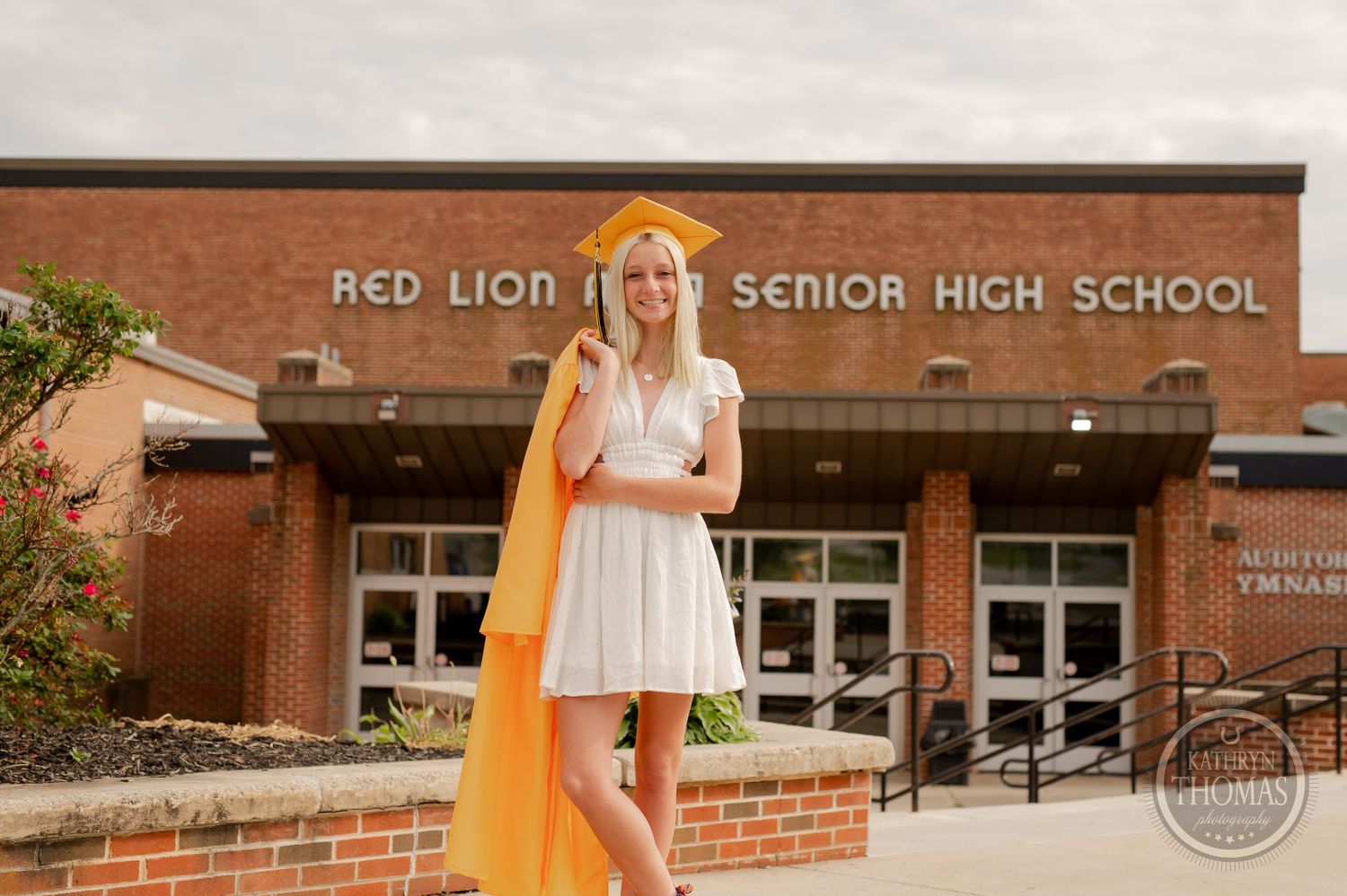 Graduate in white dress with yellow cape stands at Red Lion High School entrance.