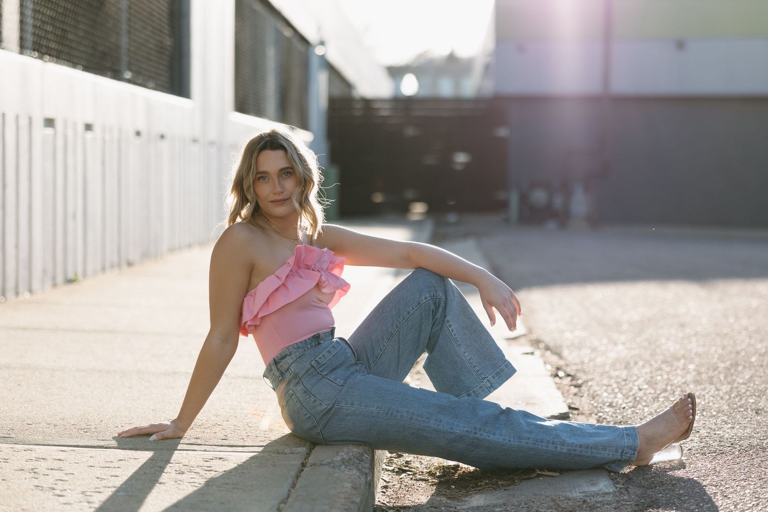Woman in pink top and jeans sitting against white wall in sunlight.