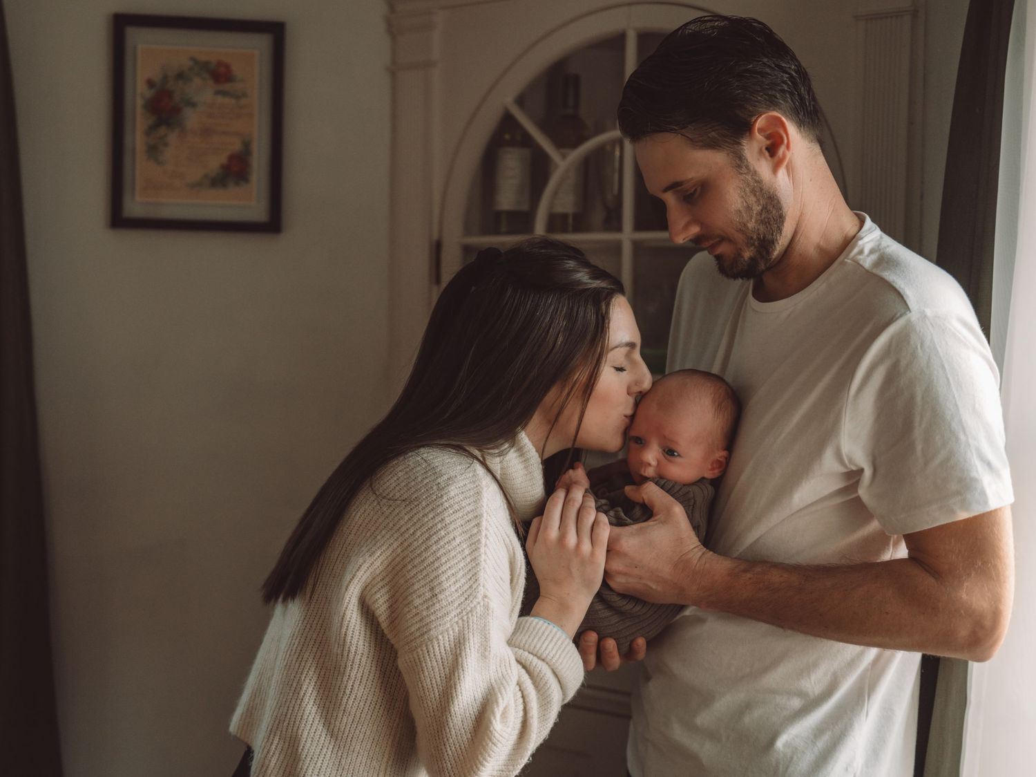 A tender family moment as parents comfort their newborn baby in a warmly lit room with white walls and a framed picture.