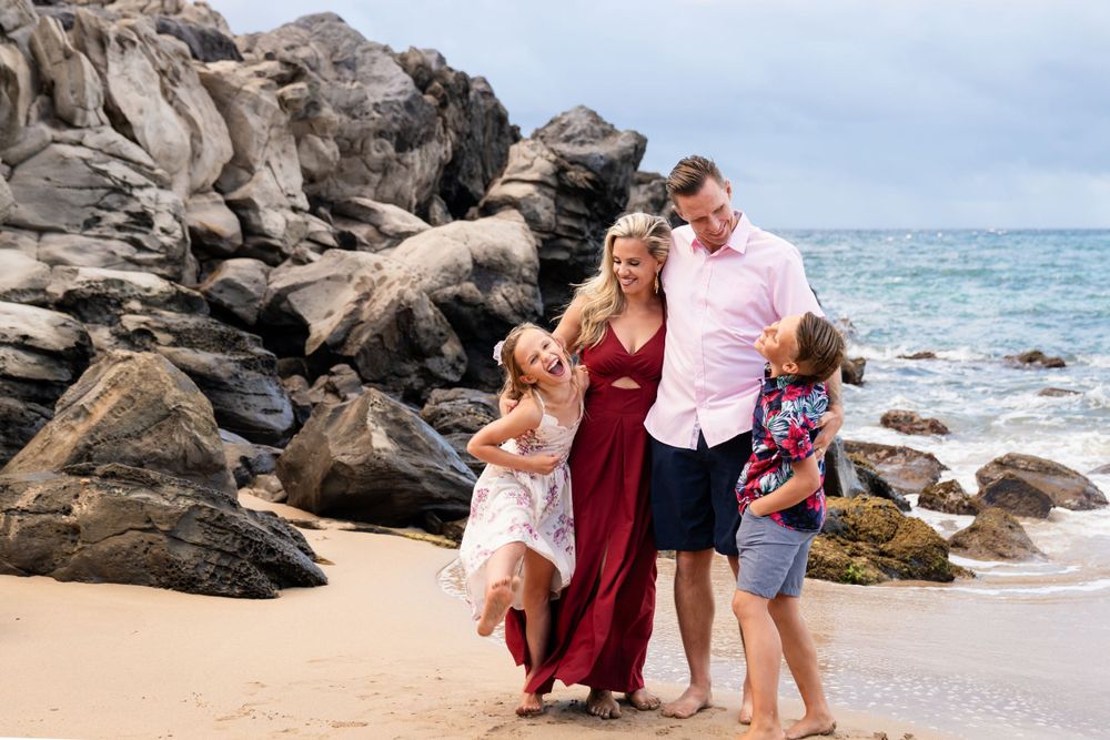 Family poses near dramatic rocky coastline with ocean waves and blue sky in background.