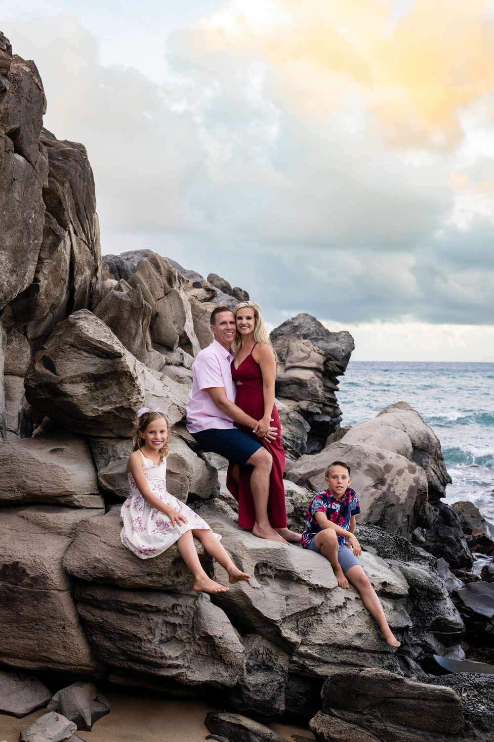 Family sitting on dramatic volcanic rocks overlooking ocean during sunset.