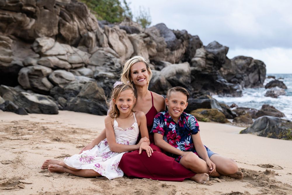 A loving family sits together on a sandy beach with rocky cliffs in the background.