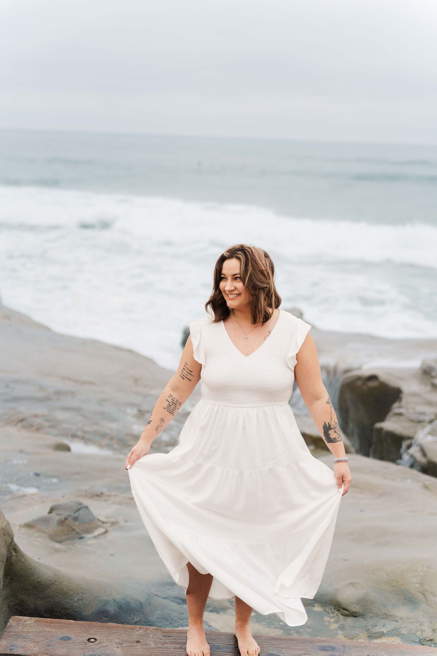 A flowing white dress catches the ocean breeze as someone stands on rocky coastal shores with waves crashing behind.