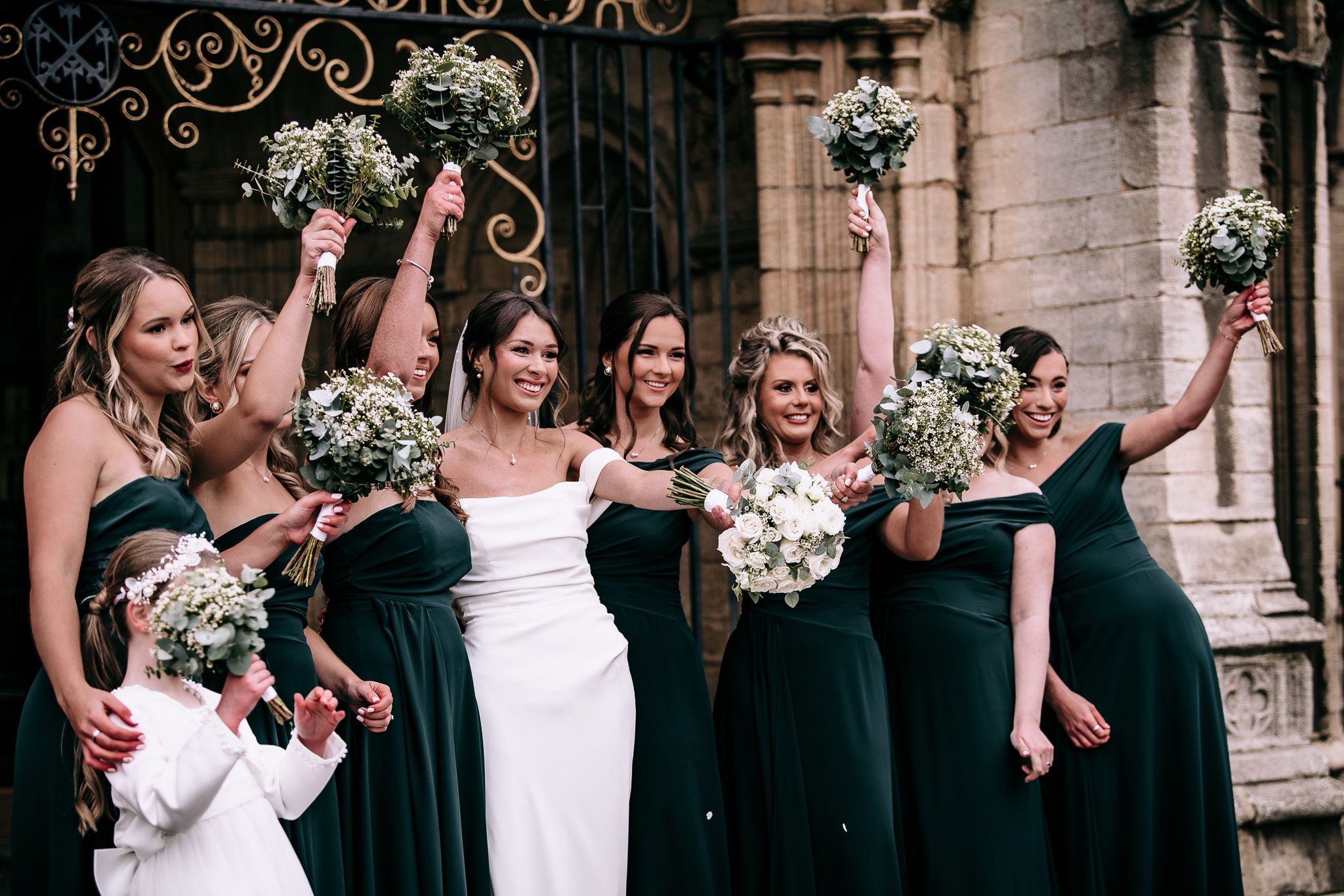 Bridesmaids in elegant dark green dresses holding floral bouquets outside a church.