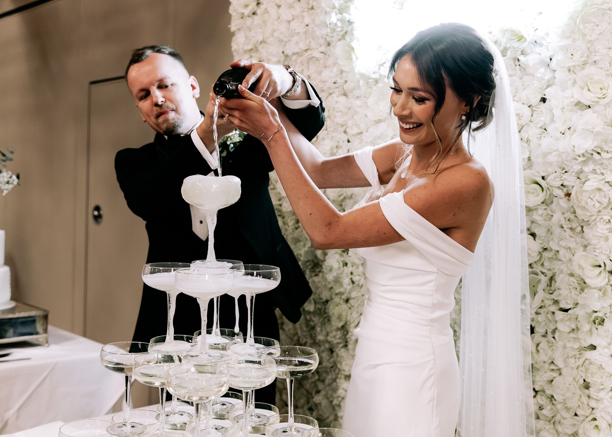 Couple pouring champagne into tower of glasses surrounded by white floral arrangements.