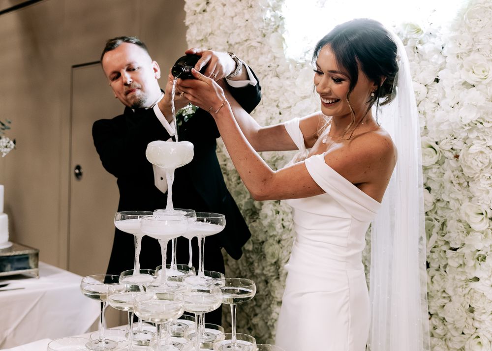 Couple pouring champagne into tower of glasses surrounded by white floral arrangements.