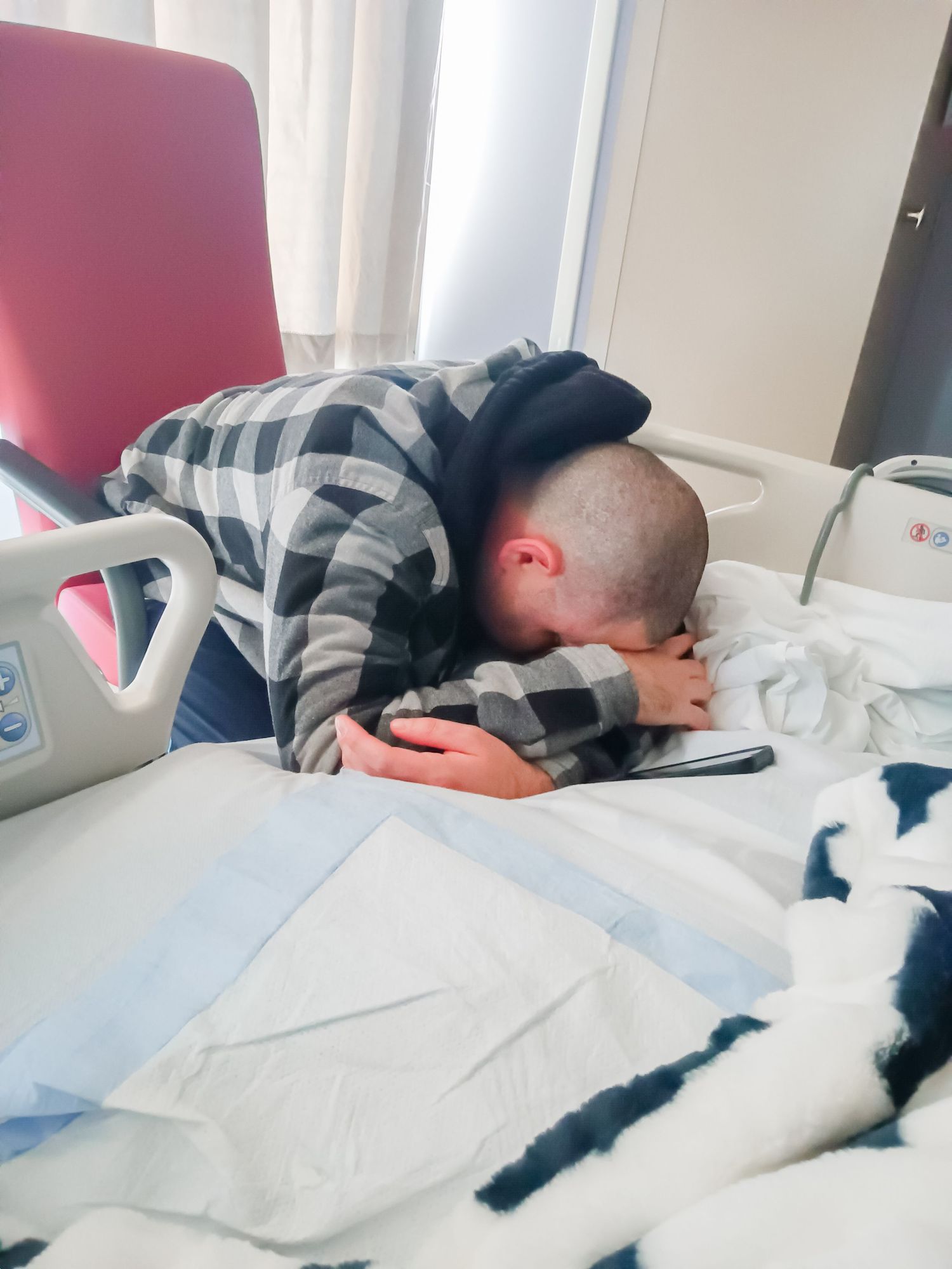 Person in plaid shirt lying face down on white bedding while looking at a device.