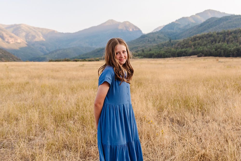 Person in blue dress stands in golden field with mountains behind.